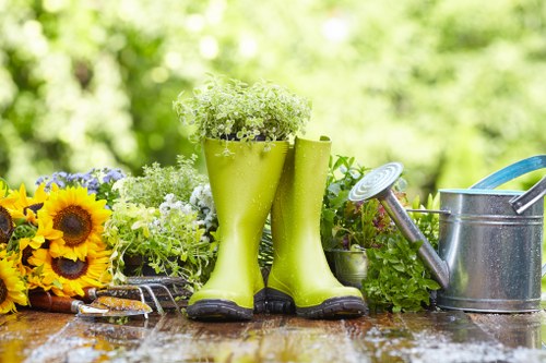 The image displays a garden scene with a wooden outdoor table featuring various gardening tools and decorative elements. In the centre, a pair of bright green rubber gardening boots are used as a planter, filled with white and green foliage plants. To the left, there is a small bouquet of vibrant yellow sunflowers and mixed flowers, adding colourful diversity to the scene. On the right, a silver metal watering can with a long spout is placed beside the boots, suggesting active gardening or watering. The background consists of a blurred view of lush green foliage and trees, indicating a well-maintained outdoor space typical of a landscaped garden at a home in Barkingside, Essex. The overall scene conveys a sense of outdoor gardening activity, seasonal growth, and the use of practical and decorative gardening accessories in a natural environment, aligning well with professional gardening and landscaping services offered by companies like Lawn Mowing Barkingside.
